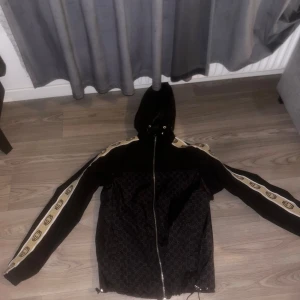 Svart Gucci vindjacka med huva - Gucci vindjacka i svart med beige och bruna GG-loggor längs ärmarna. Jackan har huva med snörning, hel dragkedja och diskret GG-mönster på framsidan. Materialet är polyester och jackan har en loose passform. Perfekt för dig som vill sticka ut med lyxig streetstyle.