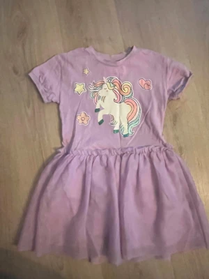 Lila enhörningsklänning från Lindex - Supergullig lila klänning från Lindex med kort ärm och ett stort tryck av en enhörning, stjärnor och blommor framtill. Kjolen är lätt utställd och klänningen är i mjuk bomull, perfekt för dig som älskar magiska motiv.