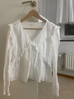 Vit blus med volang från Gina Tricot - Säljer en söt vit blus från Gina Tricot med v-ringning och långa ärmar. Helt ny, aldrig använt men bara lite skrynklig 