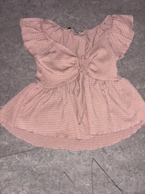 Rosa blus med volang och knyt - Supersöt rosa blus med volangärmar och knyt framtill. Blusen har en lätt rynkad struktur och en luftig, flowy passform. Perfekt för dig som gillar romantiska detaljer och en feminin look.