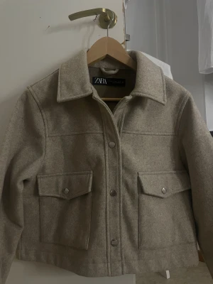 Beige overshirt från Zara - Snygg beige overshirt från Zara med klassisk krage och stora fickor framtill med tryckknappar. Jackan har en rak och loose passform, tillverkad i mjukt ullmaterial som ger en chill vibe. Perfekt att slänga över en hoodie eller t-shirt för en laidback look.