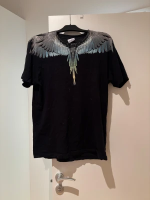 Svart t-shirt Marcelo Burlon med vingar - Svart t-shirt från Marcelo Burlon County of Milan med ikoniskt vingtryck i grått, blått och grönt över axlar och hals. Modellen är rak med rund hals och korta ärmar. Materialet är mjuk bomull som känns skön mot huden. Perfekt för dig som gillar streetwear med edge.