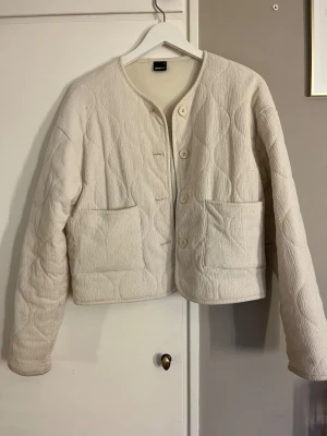 Beige quiltad jacka från Gina Tricot - Quiltad beige jacka från Gina Tricot i storlek S. Jackan har rund hals, stora framfickor och knäppning med fyra knappar. Materialet är mjukt och strukturerat, med ett vågigt quiltat mönster som ger en chill vibe.