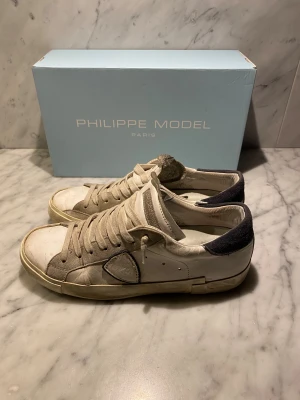 Philippe Model sneakers 41 - Snygga Philippe Low sneakers i storlek 41. Skorna är vita med beige och grå detaljer, har rund tå och platt sula. Ovandelen är i skinn och mocka, med mörkgrå häl och klassisk snörning. Perfekt för dig som gillar stilrena och exklusiva sneakers. Box ingår!