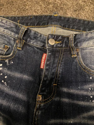 Dsquared2 med slitningar - Snygga blå jeans från Dsquared2 med skinny passform och coola slitningar samt vita färgstänk. Klassisk femficksmodell med gul kontrastsöm och svart läderpatch bak. Perfekt för dig som gillar streetstyle och detaljerad denim.