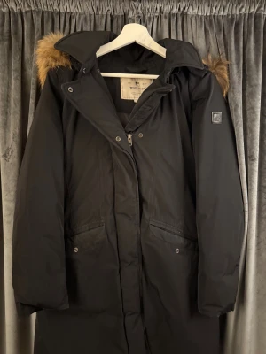 Svart parkas från Woolrich med pälskrage - Säljer en svart parkas från Woolrich med snygg pälskant på huvan. Jackan har dragkedja och tryckknappar framtill, två stora fickor och en klassisk patch på ärmen. Perfekt för kalla dagar och riktigt stilren look. Orginal pris 7000kr