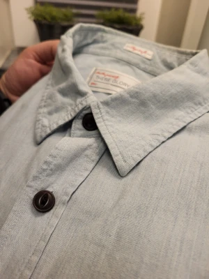 Ljusblå skjorta från These Glory Days - Ljusblå skjorta från These Glory Days med klassisk krage, svarta knappar och två bröstfickor med lock. Skjortan har axelklaffar och är långärmad. Materialet är mjukt och känns som bomull, perfekt för en chill och stilren look.