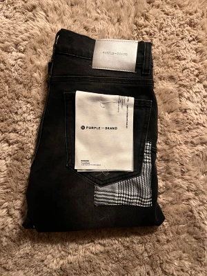 Svarta jeans från Purple Brand - Svarta jeans från Purple Brand med slim fit passform och slitna detaljer på benen. Baksidan har en unik rutig patch på fickan och silverfärgad logotyp på linningen. Jeansen är gjorda i stretchigt bomullsmaterial och har klassisk femficksdesign. (Pris går att diskuteras)