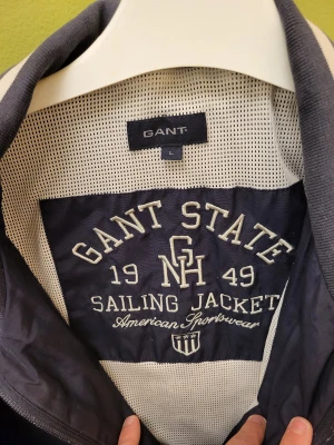 Marinblå vindjacka från Gant - Snygg marinblå vindjacka från Gant med broderad logga och text på bröstet och insidan. Jackan har dragkedja, ribbade muddar och krage med vit rand. Tillverkad i polyester med meshfoder för extra komfort. Perfekt för dig som gillar sportig och klassisk stil.