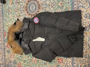 Svart dunjacka från Canada Goose  - Säljer en svart dunjacka från Canada Goose med ikonisk patch på ärmen. Jackan har huva med snygg pälsdetalj, långa ärmar och stängs med både dragkedja och knappar. Perfekt för kalla vinterdagar och riktigt varm. Klassisk och stilren design.
