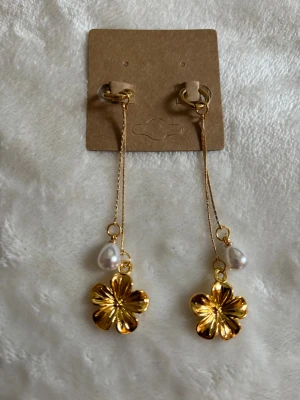 Guldiga örhängen med blomma och pärla - Snygga örhängen i guldfärgad metall med hängande blommor och små vita pärlor. Kedjorna är tunna och ger ett elegant intryck. Perfekta för att lyfta vilken outfit som helst med sin blommiga och glansiga look.