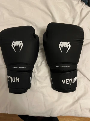 Boxningshandskar Venum 10oz - Säljer ett par svarta boxningshandskar från Venum, 10oz. Handskarna har en stilren design med vit Venum-logga och ormsymbol på ovansidan. Tillverkade i syntetmaterial med bred kardborrestängning för extra stöd runt handleden. Perfekta för träning och sparring.