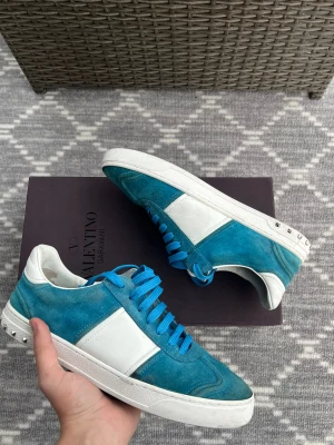 Valentino Garavani blå sneakers - Snygga sneakers från Valentino Garavani i blå mocka med vita detaljer och blå snörning. Skorna har platt sula och silverfärgade nitar på hälen för en cool touch. Perfekt för dig som vill sticka ut med stil. Skriv vid eventuella.