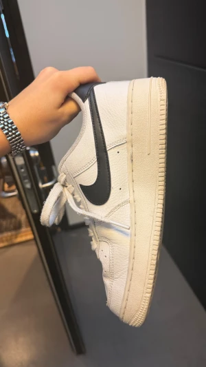 Nike Air Force 1 vita och svarta sneakers - Säljer ett par klassiska Nike Air Force 1 sneakers i vitt skinn med svart swoosh och häl. Använda typ 5 gånger. En liten tvätt så ser dem ut som helt nya. Defekt: sulan har kasat fram lite och en tråd vid sulan/ sömmen sticker ut. Skorna har snörning och är perfekta för dig som gillar streetwear och tidlös stil.