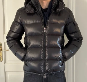 Moncler Maya - Säljer en svart pufferjacka från Moncler med glansig finish, huva och praktisk ficka med logotyp på ärmen. Jackan har dragkedja framtill och är vadderad för extra värme. Perfekt för dig som vill ha en snygg och ikonisk vinterjacka.