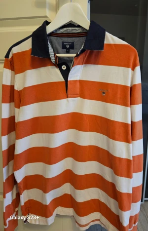 GANT orange/vit pikétröja, storlek L - Randig långärmad pikétröja med en mörkblå krage och en broderad logotyp på bröstet.