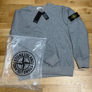 Stone island Sweatshirt - Snygg grå sweatshirt från Stone Island med klassisk rund hals och ribbade muddar. Ikonisk svart och gul logotyp-patch på vänster ärm. Mjuk bomullskvalitet som känns skön mot huden. Perfekt för dig som gillar streetwear och vill ha en clean look. TVEKA INTE ATT SKICKA EN BUD:)