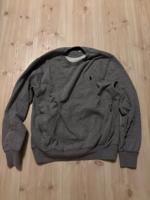 Grå sweatshirt från Ralph Lauren - Säljer en klassisk grå sweatshirt från Ralph Lauren med rund hals och broderad logga på bröstet. Tröjan har långa ärmar och är riktigt skön och enkel att matcha till olika outfits.