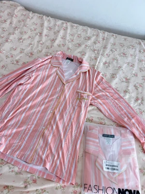 Randig rosa pyjamasskjorta från Fashionnova - Säljer en HELT NY suuuper skön glittrig långärmad pyjamas skjorta från Fashion Nova i rosa med vita och guldiga ränder. Skjortan har klassisk krage, knäppning framtill, bröstficka med broderad text och snygga guldiga detaljer längs kanterna. Materialet är otroligt skönt och skjortan är helt ny och aldrig använd. Öppnade paketet idag. 