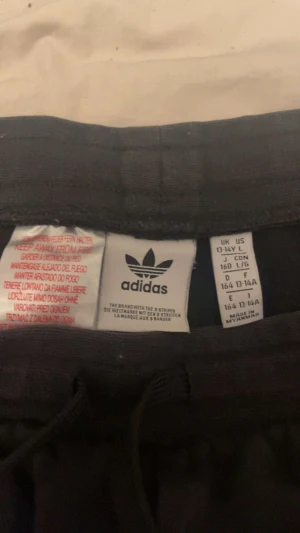 Svarta Adidas shorts med vita ränder - Svarta Adidas shorts med klassiska vita ränder längs sidorna och Adidas-logga framtill. Modellen har snörning i midjan och är tillverkad i mjukt material, perfekt för en sportig och avslappnad stil.
