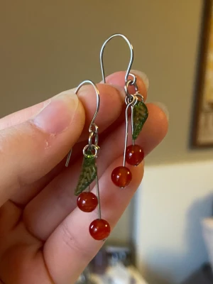 Hängande örhängen med körsbärsdetaljer - Snygga örhängen i silverfärgad metall med röda pärlor som ser ut som körsbär och gröna blad i plast. De har en lekfull och trendig design som verkligen poppar. Perfekta för dig som vill ha något unikt och färgglatt till din stil.