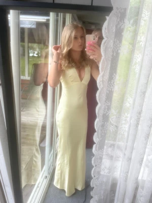 balklänning - Säljer en elegant ljusgul långklänning med djup v-ringning och figurnära passform. Klänningen är ärmlös och har en mjuk, slät yta som ger ett lyxigt intryck. Perfekt för dig som vill sticka ut med en stilren och feminin look. använd en gång