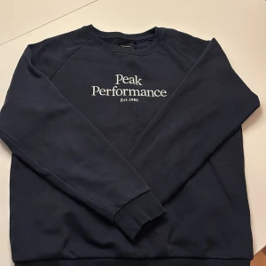 Mörkblå Peak Performance sweatshirt - Snygg mörkblå sweatshirt från Peak Performance med vit broderad logga på bröstet. Tröjan har rund hals, ribbade muddar och är tillverkad i mjuk bomullsmix. Perfekt för chill eller sportiga dagar.