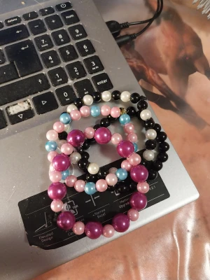 Färgglada pärlhalsband och armband - Säljer ett set med pärlhalsband och armband i plast med stora och små pärlor i lila, rosa, blått, svart och vitt. Perfekt för dig som gillar färg och vill sticka ut med din stil. Pärlorna är runda och har en glansig finish.