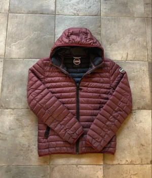 Mörkröd Colmar pufferjacka med huva - Snygg mörkröd pufferjacka från Colmar med huva och svarta dragkedjor. Jackan har quiltad design, långärmade ärmar och logotyp på vänster arm. Insidan är mörkblå och jackan har två sidofickor med dragkedja. Perfekt för kyliga dagar.