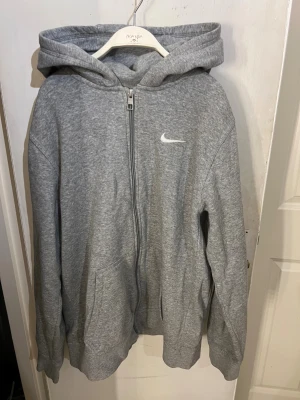 Grå hoodie från Nike med dragkedja - Grå hoodie från Nike med vit logga på bröstet. Tröjan har huva, långa ärmar och praktiska fickor framtill. Materialet är mjuk bomull och polyester, perfekt för chill eller träning. Det står storlek XL i tröjan men den sitter med som en storlek S/M