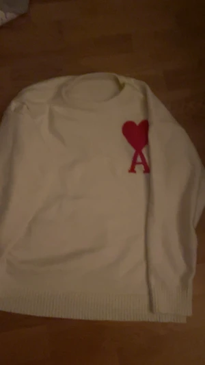 Vit stickad tröja med rött hjärta - Säljer en vit stickad tröja från AMI Paris med ett stort rött hjärta och bokstaven A broderat på bröstet. Tröjan har rund hals och ribbade muddar vid ärmslut och nederkant. Perfekt för dig som gillar stilrena detaljer och vill sticka ut.
