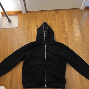 Svart hoodie från Burberry passar m också - Svart hoodie från Burberry i storlek L med klassiskt rutigt foder i huvan. Tröjan har dragkedja framtill, broderad logga på bröstet och två stora fickor. Perfekt för dig som vill ha en stilren och lyxig streetwear-look.