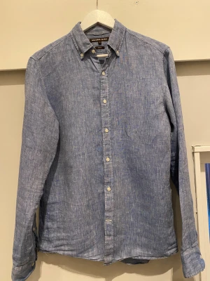 Blå linneskjorta Michael Kors S - Snygg blå linneskjorta från Michael Kors i slim fit-modell. Skjortan har klassisk krage med knappar, långa ärmar och diskret melerat mönster. Perfekt för dig som gillar stilrena och bekväma plagg med en modern touch. (Pris kan diskuteras vid smidig affär)