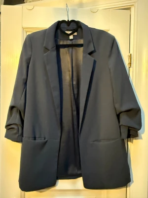 Mörkblå kavaj med ”uppdragna” ärmar, se bild - Snygg mörkblå kavaj med klassisk krage och uppvikta ärmar. Modellen är rak och har två stora fickor framtill. Perfekt för en stilren look och enkel att matcha med olika outfits.