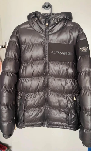 Svart pufferjacka Alessandro Zavetti - Snygg svart pufferjacka från Alessandro Zavetti med glansig finish, huva med snörning och dragkedja framtill. Jackan har två fickor med dragkedja och logga på bröstet samt ärmen. Perfekt för kalla dagar och riktigt stilren look.