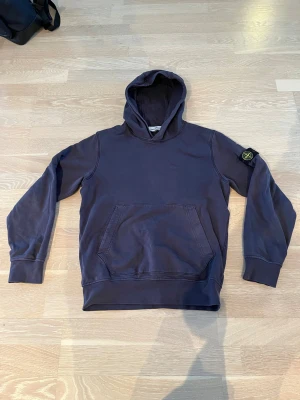Mörkblå Stone Island hoodie 13år - Snygg mörkblå hoodie från Stone Island Junior med klassisk logotyp-patch på vänster ärm. Tröjan har huva, magficka och ribbade muddar. Tillverkad i mjuk bomull, perfekt för chill dagar. Enkelt och stilrent plagg som ger street-vibe. 