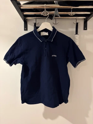 Marinblå pikétröja från Ami Paris - Snygg marinblå pikétröja från Ami Paris med vit piping på krage och ärmslut. Klassisk polokrage med två knappar och diskret Ami Paris-brodyr på bröstet. Tillverkad i mjuk bomull, perfekt för en chill och stilren look. Nypris: 1645kr