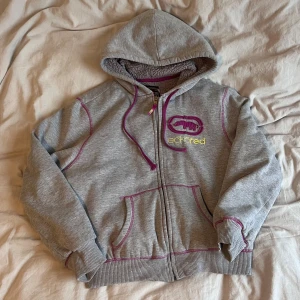 Grå hoodie från Ecko Red med lila detaljer - Snygg grå hoodie med lila sömmar och snören från Ecko Red med dragkedja, lila broderad logga och gula detaljer på bröstet samt fodrad på insidan. Väldigt varm och mysig! Köpt second hand och har endast använts av mig ett fåtal gånger