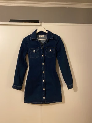 Mörkblå jeansklänning från Zara - Snygg kort mörkblå jeansklänning från Zara authentic denim by TRF med långa ärmar,  krage och knappar hela vägen framtill. Klänningen har två bröstfickor med lock och silverfärgade knappar. 