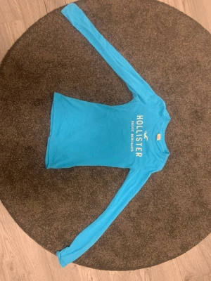 Ljusblå långärmad tröja från Hollister - Snygg ljusblå långärmad tröja från Hollister med vit logotyp och text på bröstet. Tröjan har en slim passform och är perfekt för dig som gillar en enkel och fräsch stil. Rund hals och klassisk design. Hör av dig vid frågor💕