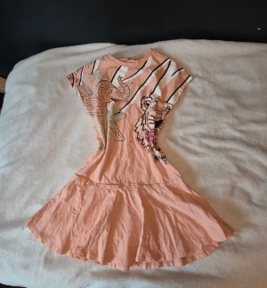 Peachfärgad Kenzo klänning med djurmotiv - Superfin peachfärgad kortklänning från Kenzo med coolt tryck av elefant och tiger både fram och bak. Klänningen har rund hals, korta ärmar och är gjord i mjuk bomull. Perfekt för dig som gillar unika prints och streetstyle vibes.