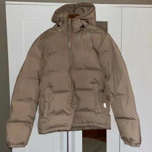 Beige pufferjacka med huva - Snygg beige pufferjacka med huva och dragkedja framtill, från Jack&Jones i storlek M. Jackan har en clean look, två fickor inne i jackan och är vadderad för extra värme. Perfekt för kalla dagar och enkel att styla till olika outfits. Perfekt för julklapp till både killar och tjejer😍