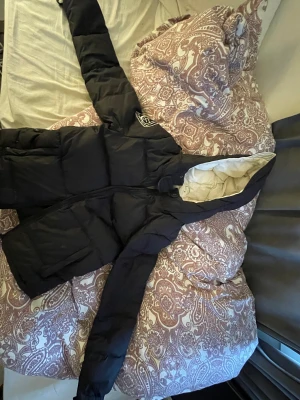 Svart pufferjacka från db, stl S - Svart pufferjacka från db med broderad logga på ärmen och huva. Jackan har en quiltad design och dragkedja framtill. Insidan är ljus och jackan har långa ärmar. Perfekt för kyliga dagar.