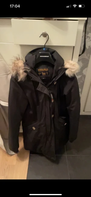  Woolrich jacka med päls - Svart parkas från Woolrich med stor huva och fluffig beige päls.. Jackan har dragkedja och knappar framtill, praktiska fickor och en klassisk, passform. Perfekt för kalla dagar och har en snygg streetstil. 