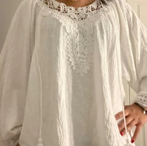 Vit boho-blus med spetsdetaljer - Supersöt vit blus i boho-stil med breda långärmar och snygga spetsdetaljer vid halsringningen och ärmsluten. Blusen har en luftig passform och är gjord i ett lätt, krinklat tyg med dekorativa snören framtill. Perfekt för dig som gillar romantiska och trendiga plagg.