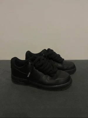 Svarta Nike Air Force 1 med rep snören - Säljer ett par helsvarta Nike Air Force 1 med extra tjocka svarta snören. Skorna har klassisk rund tå, perforerad tåbox och är tillverkade i skinn. Perfekta för dig som vill ha en clean och stilren sneaker med en unik twist.