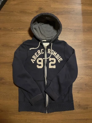 Mörkblå Abercrombie Zip hoodie - Riktigt snygg abercrombie & fitch zip hoodie i storlek L/XL, står XL inne i tröjan men jag har oftast L och den passar mig bra. Ganska slim passform överlag. Släng gjärna iväg en fråga om det behövs!           OBS: varan har fabriktillverkade slitningar!!