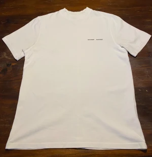 Vit t-shirt från Samsøe Samsøe - Vit t-shirt från Samsøe Samsøe med minimalistisk svart logotyp på bröstet som endast är använd några få gånger. Modellen har en högre rund hals och raka korta ärmar. Materialet är mjuk bomull och passformen är normal. Perfekt för en clean och stilren look. (Pris går att diskutera vid smidig affär)