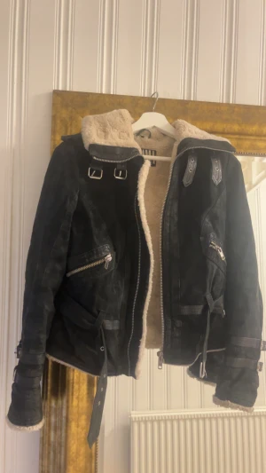 Svart teddyjacka från Jofama by Kenza - Säljer en svart teddyjacka från Jofama by Kenza med beige fluffigt foder. Jackan har coola detaljer som spännen vid kragen, dragkedjor på fickorna och bälte vid midjan. Perfekt för dig som vill ha en snygg och varm jacka med edge. Jackan är i använt skick men endast legat i garderoben senaste åren. 