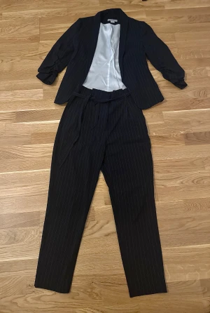 Svart randig kostym set från H&M, strl 34 - Säljer en svart kostym från H&M i storlek 34. Setet består av en kavaj med upprullade ärmar och matchande byxor med knytband i midjan. Tunt vitt foder i kavajen och smala vita ränder över hela kostymen. Perfekt för dig som gillar stilrena och klassiska vibes.